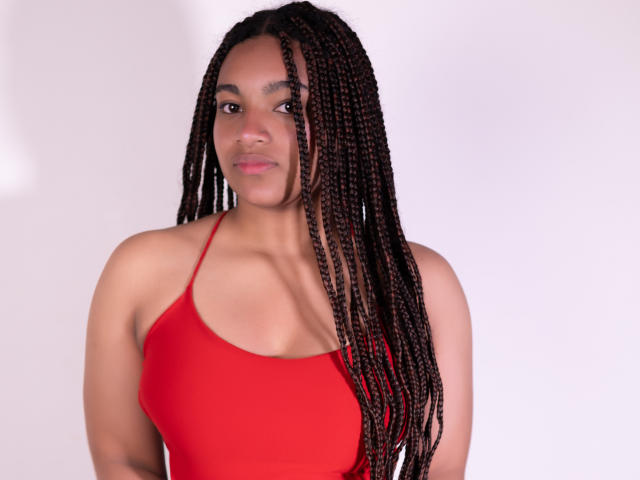 KathePalmer, avec ses longs cheveux bruns tressés, porte un haut rouge à bretelles fines qui révèle ses petites seins fermes, et elle sourit discrètement à l'appareil photo..