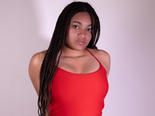 KathePalmer, avec de longs cheveux tressés, portant un débardeur rouge, montrant de petites seins, une peau brune et une expression neutre, debout devant un fond blanc uni..