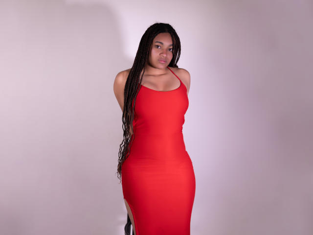 KathePalmer, vêtue d'une robe rouge moulante, debout les bras derrière le dos, avec de longs cheveux tressés, de taille moyenne, avec de petites seins, regardant la caméra, peau claire, expression neutre..