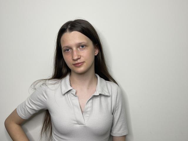 Joetta Savka tem cabelos castanhos longos, pele clara e seios pequenos. Ela está vestindo uma camisa polo branca, em pé em frente a uma parede branca lisa, com um sorriso discreto..
