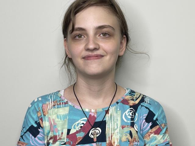 GiannaEget, con capelli castani, pelle chiara, seno piccolo, indossa una camicia colorata con un motivo astratto e una collana nera con un ciondolo circolare, sorridendo dolcemente..