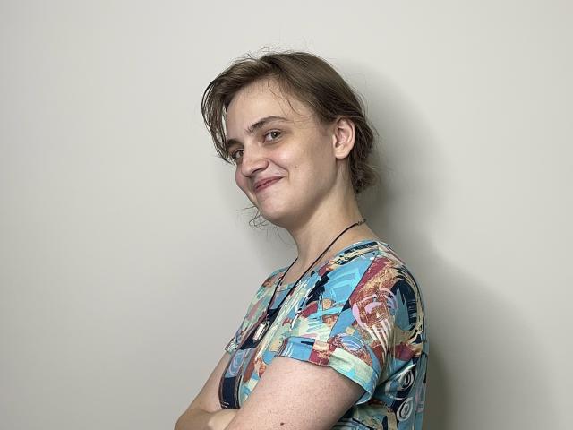 GiannaEget with messy brown hair, smiling slightly, wearing a colorful patterned shirt, arms crossed, necklace visible, white wall background, casual pose.