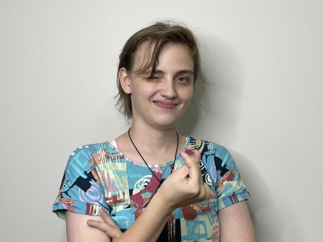 GiannaEget with a slight smile, brown hair, and a colorful geometric shirt, pointing at her chest with a finger, wearing a black necklace against a plain white wall.