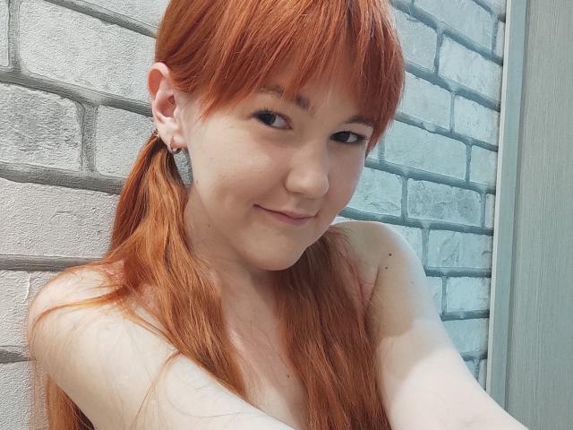 SheritaChieng a les cheveux roux en couettes, de petites seins, une peau claire et un sourire mignon. Elle porte des boucles d'oreilles et un petit anneau au nez. Elle prend un selfie..