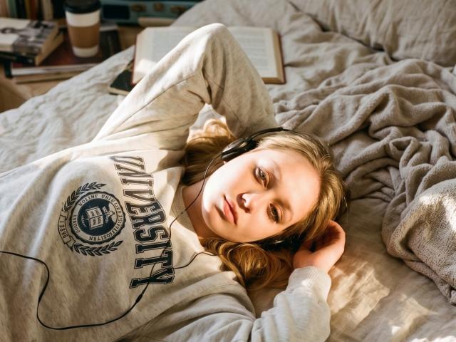 CherSartell lies on a bed, wearing headphones, with a gray "UMICH" hoodie, her blonde hair spread, eyes half-open, looking relaxed, arm behind head, soft sunlight on her face.