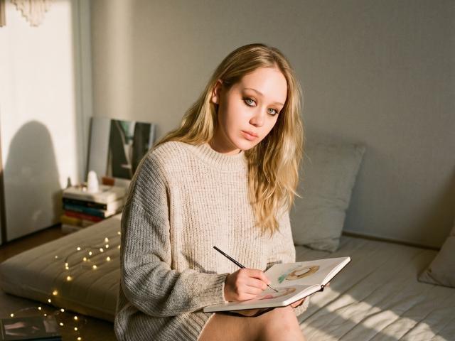 CherSartell with blonde hair, wearing a beige knit sweater, sitting on a bed, writing in a notebook with a pencil, looking down, sunlight casting shadows.