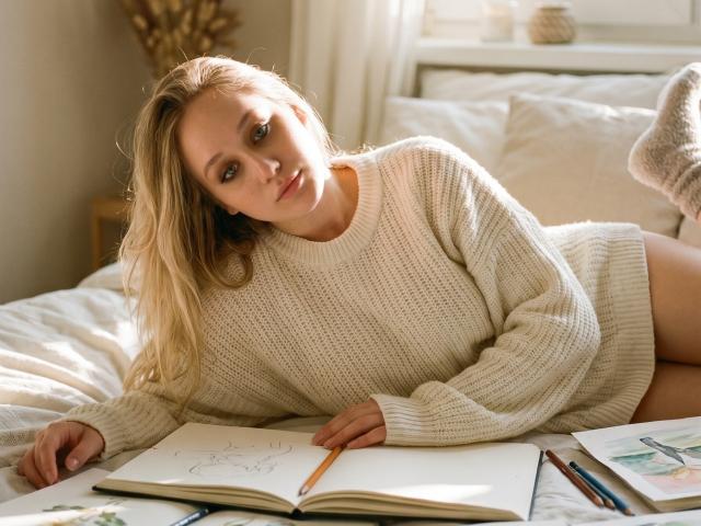CherSartell with blonde hair, lying on bed, wearing a beige knit sweater, holding pencil in sketchbook, pens on bed, soft light, slightly tilted head, relaxed, attractive.