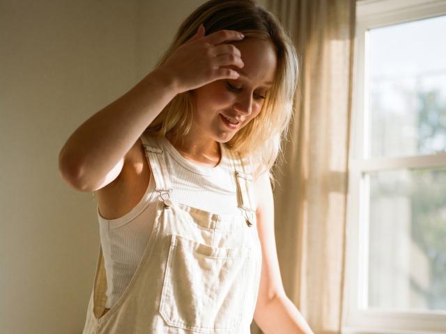 CherSartell with blonde hair, wearing white overalls and a white shirt, touches her head, smiling softly, sunlight through a window.