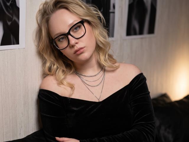 CherSartell with blonde wavy hair, black glasses, off-shoulder black velvet top, and silver chains, looking at camera with a neutral expression, arms crossed.