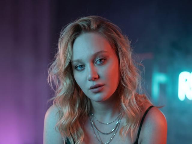 CherSartell has wavy blonde hair, blue eyes, and a serious expression. She's wearing a black top and multiple necklaces, with red and blue lighting on her face.
