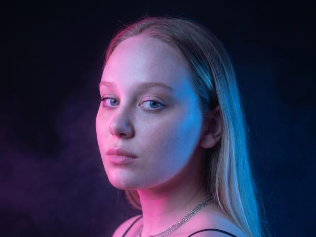 CherSartell's face, lit with pink and blue light, showing pale skin, blonde hair, subtle makeup, and a silver necklace, looking slightly to the side with a neutral expression.