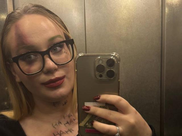 CherSartell with blonde hair, black glasses, red lipstick, and tattoo on neck, holding iPhone in elevator, wearing black top, red nails, ring on finger.