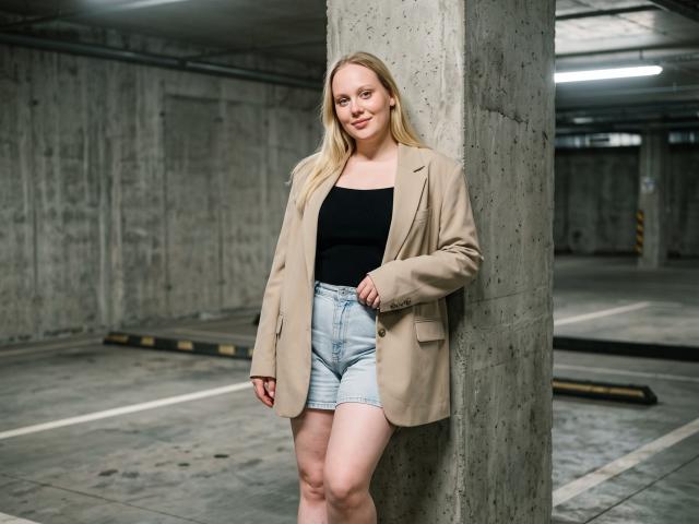 CherSartell with blonde hair, black top, beige blazer, and light blue denim shorts, standing against a concrete pillar in a parking garage, smiling slightly.