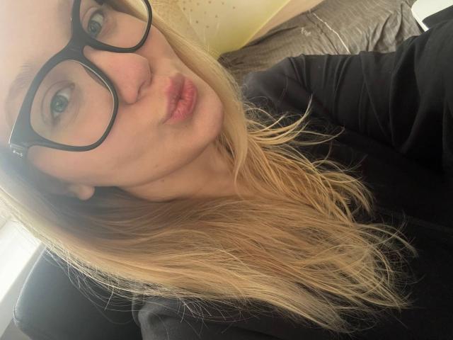 CherSartell, avec des lèvres boudeuses, des cheveux blonds, des lunettes noires et un haut noir, regardant la caméra, montrant son nez et ses lèvres légèrement entrouvertes, roses. Ses seins ne sont pas visibles..