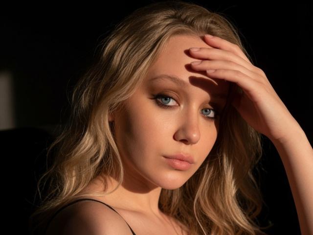 CherSartell has blonde wavy hair, blue eyes, and a hand on her head, looking slightly to the side with a serious expression, wearing a black strap.