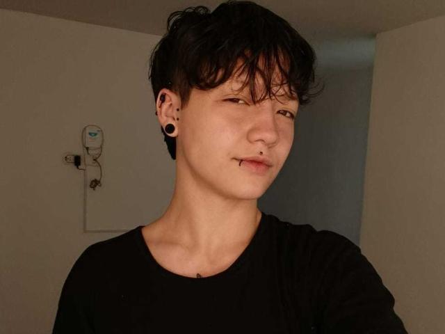 LissyLuxe, short black hair, black ear stretchers, black shirt, nose ring, slight smirk, light skin, subtle makeup, visible scar below right eye, dimly lit room.