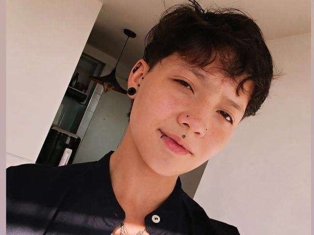 LissyLuxe has short, messy black hair, a nose piercing, and a lip piercing with black ear plugs. She's wearing a black shirt, smiling softly, and has a cute face.