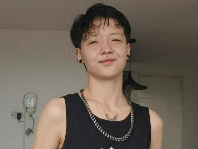 LissyLuxe, a skinny dude with short black hair, wearing a black tank top and chain necklace, smiling with small black ear plugs and a tattoo on his chest.