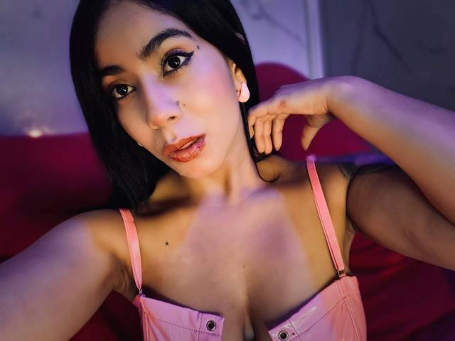 MarianaJackson with black hair, pink lingerie, and glossy lips, showing off her tits and a tattoo on her arm, with her hand near her face.