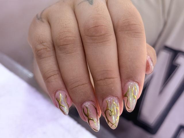 NatalieHarris' hands with long, glittery nails featuring gold and pink patterns, her fingers slightly curled, showing a small tattoo on her left index finger.