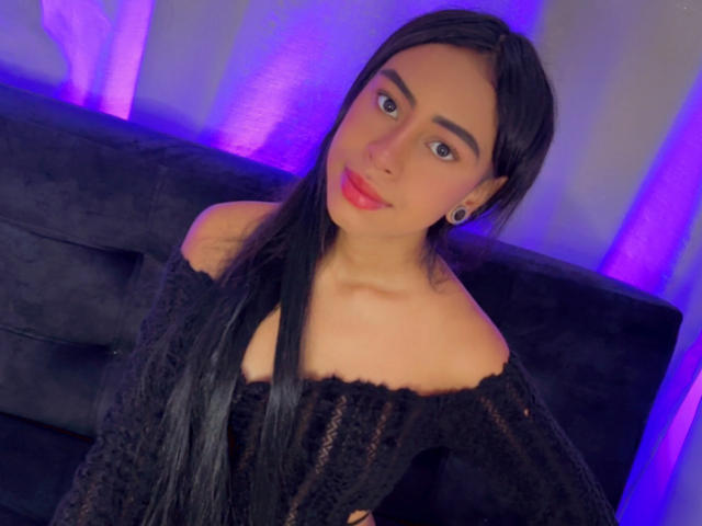 SiennaVance with long black hair, wearing an off-shoulder black lace top, sitting on a black couch, blue-purple lights in the background, and small hoop earrings.