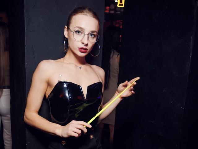 PetrinaRaven with glasses, red lipstick, and hoop earrings, wearing a shiny black strapless top, holding a yellow stick, standing in a dark, narrow space.