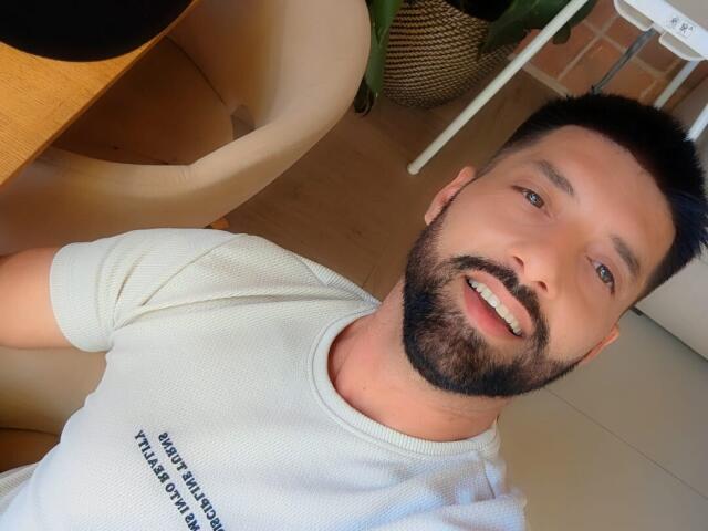 TommyDum, with a short, dark beard and black hair, wearing a white t-shirt, smiling at the camera, has a cute, sexy face and a medium build.