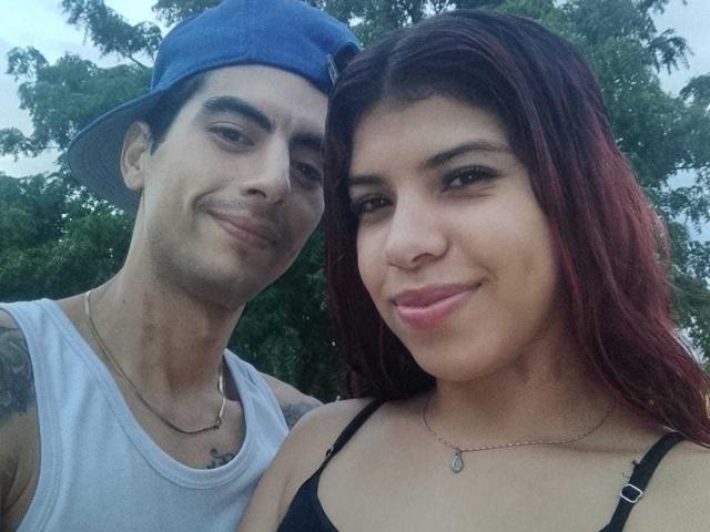 WildloversOne with dark red hair and pink lips, wearing a black bra, next to WildloversOne in a blue cap and white tank top, both smiling.