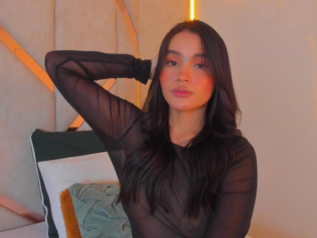 MiiaPalmer with long black hair, wearing a sheer black long-sleeve top, has her right arm raised, showing her tits, and a slight pink blush on her face.