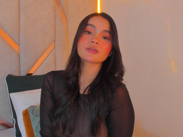 MiiaPalmer with long black hair, wearing a sheer black top, has red cheeks, and a slightly open mouth, looking at the camera with a sexy, confident expression.