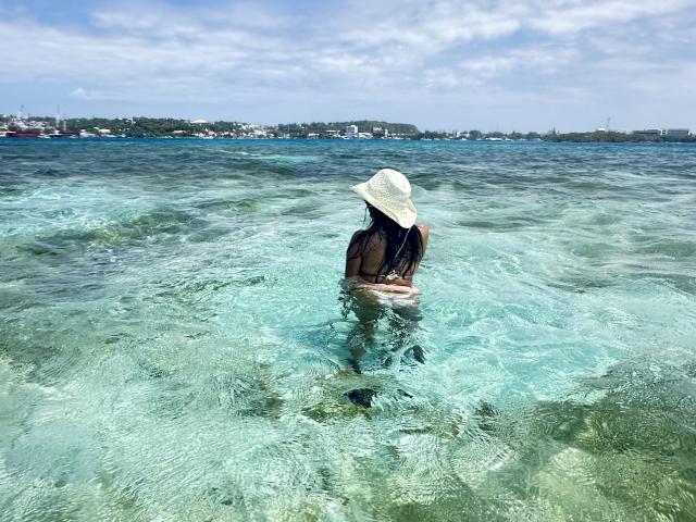 GraceClarke's ass and back are in clear blue water, wearing a white hat, with her dark hair flowing. She's standing with her legs apart, facing away.