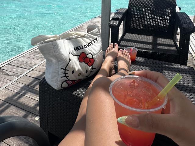 GraceClarke's legs on a wicker chair by a blue pool, holding a pink drink with a yellow straw, white Hello Kitty bag, and her feet up, black chair nearby.