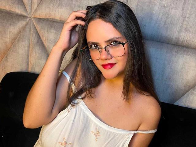 Emily Colsson, met een bril, rode lippenstift en bruin haar, draagt een witte top met een open schouder en raakt haar haar aan. Haar borsten zijn zichtbaar. Ze heeft een zachte glimlach. Lichte huid, schattig..