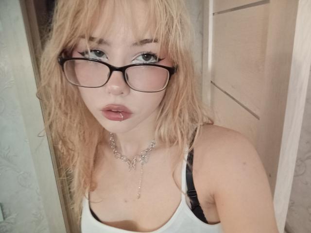 Blonde EileneSzala with glasses, lip ring, white tank top, black strap, and silver necklace, showing her tits and ass in a selfie, standing near a door.