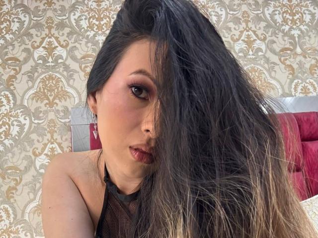 HannaRobertss has long, messy black and blonde hair covering one eye, wearing a black mesh top, with pink eye makeup and red lipstick, looking seductively at the camera.
