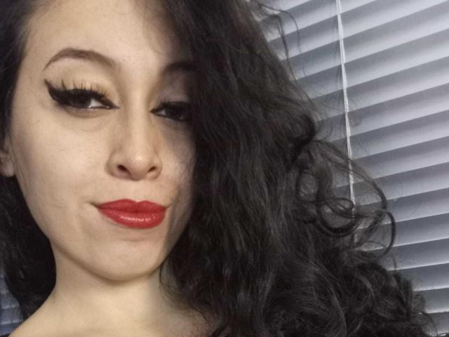 KendraSimons with long black wavy hair, red lipstick, and thick black eyelashes, smiling slightly. Her white blinds background is blurred. Boobs not visible.