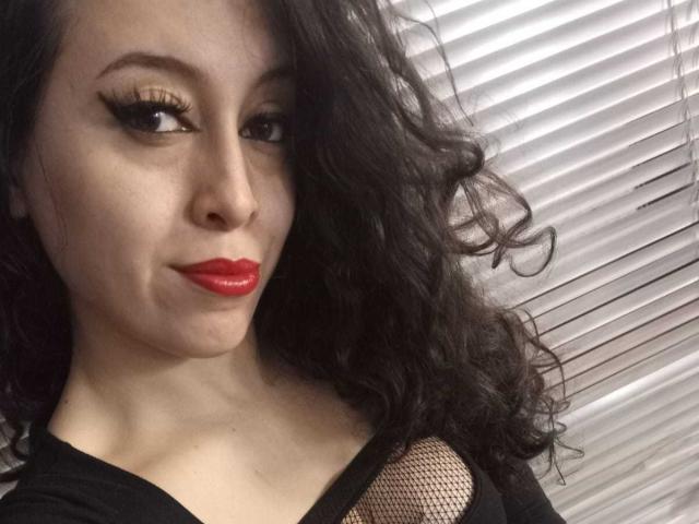 KendraSimons with curly black hair, red lipstick, and black eyeliner, wearing a black top with fishnet detail, showing her tits slightly. She's smiling at the camera.