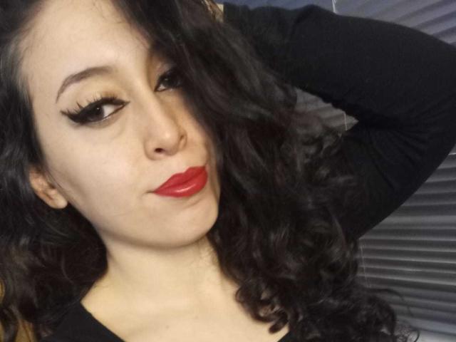 KendraSimons with dark wavy hair, red lipstick, and black eyeliner, wearing a black long-sleeve top, has her arm raised, looking slightly to the side with a subtle smile.