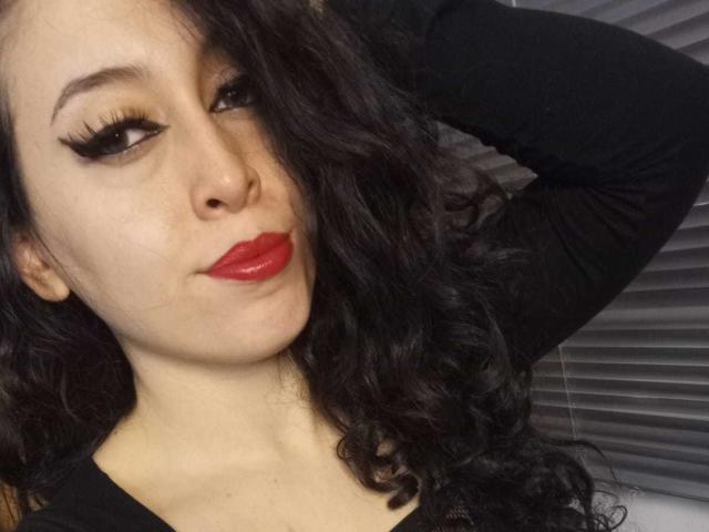 KendraSimons with curly black hair, bold red lipstick, and thick black eyeliner, wearing a black long-sleeve shirt, arm raised, looking at camera with a slight smirk.