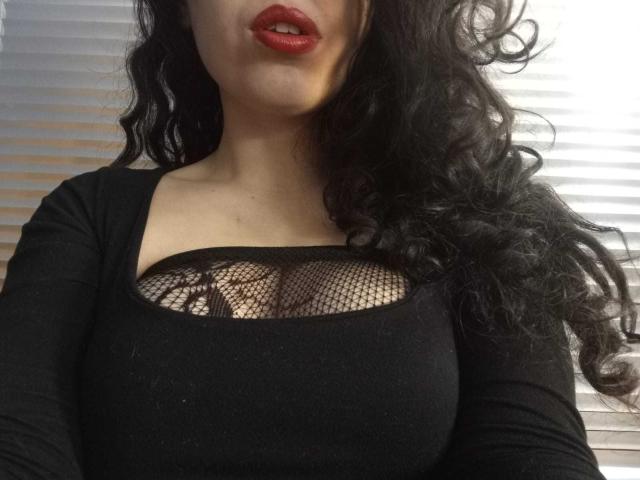 KendraSimons with long curly black hair, red lipstick, wearing a black top with a mesh cutout, showing her big boobs and black lace bra underneath.