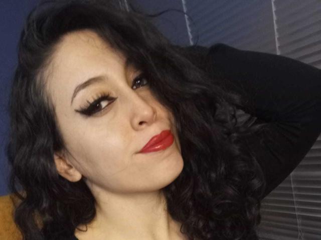 KendraSimons with black wavy hair, red lipstick, and thick black eyeliner, raising her right arm behind her head, looking slightly to the side with a subtle smirk.