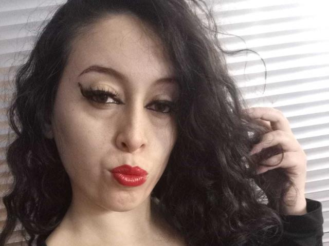 KendraSimons with dark wavy hair, red glossy lips, and thick black eyeliner, puckering her lips slightly, touching her hair with a right hand.