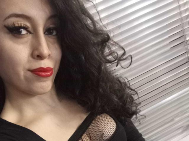 KendraSimons with curly black hair, red lipstick, and thick eyeliner, wearing a black top with mesh, looking to the side, showing her tits.