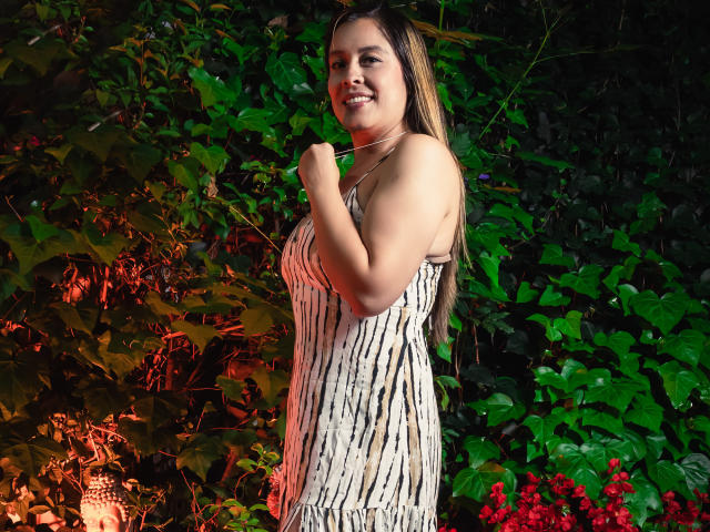 BellaMilfkm, com longos cabelos castanhos, sorrindo, vestindo um vestido branco listrado, em pé em um jardim verde e cheio de folhagens, segurando um colar, com um lanterna e flores vermelhas ao fundo..