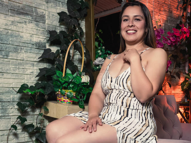BellaMilfkm com seios médios, sorrindo, vestindo um vestido listrado, sentada em uma cadeira rosa, com a mão no peito, cabelo castanho, plantas verdes e uma parede de tijolos ao fundo..