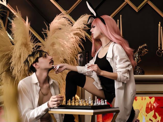 GayaAndChaim with pink hair and white bunny ears, wearing a black top and white shirt, sits on a table, showing her tits, while Chaim, in a white shirt, kneels.