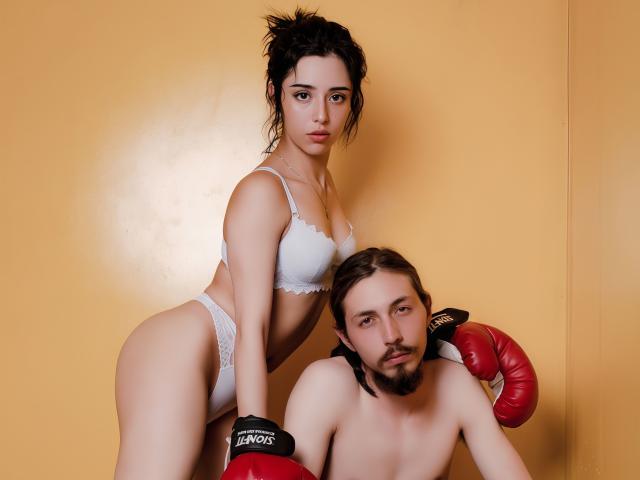 GayaAndChaim with tits in white lingerie, standing behind Chaim, who's shirtless with red boxing gloves, in a yellow-walled room. Chaim has a beard and short brown hair.