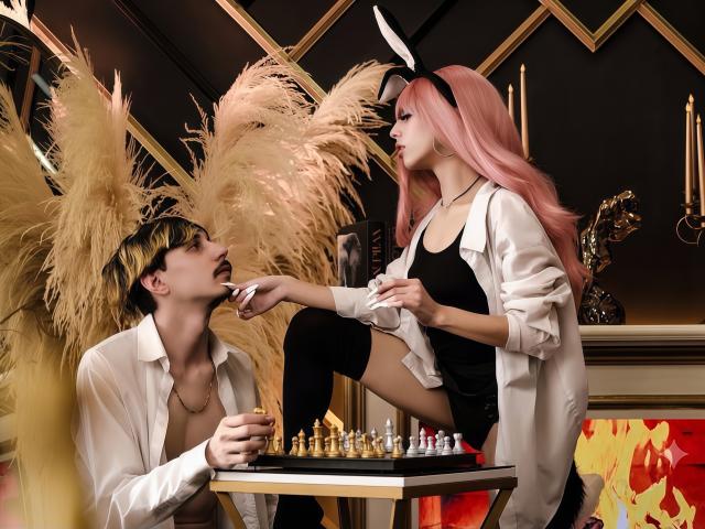 GayaAndChaim with pink hair and white bunny ears, wearing a black top and white shirt, sits on a table, extending her hand to GayaAndChaim's mouth, who's playing chess.