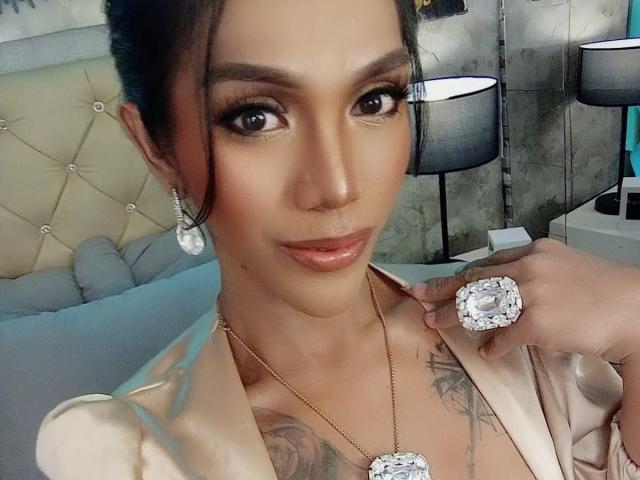 XTREMELYBeautyEmpire con labios marrones brillantes, cejas oscuras y ojos grandes, vistiendo un collar de oro y un anillo de diamantes, mostrando un tatuaje en el pecho, con una blusa de satén..