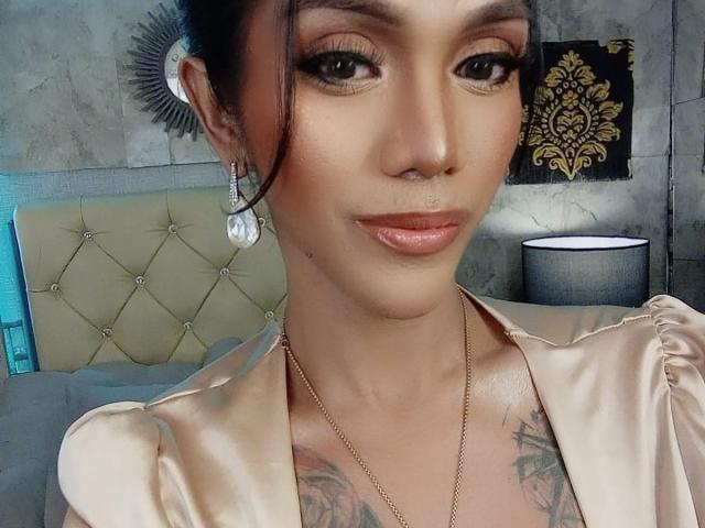 XTREMELYBeautyEmpire with shiny lips, brown eyes, and tattoos, wearing a silky beige top, gold necklace, and dangling earrings, smiling at the camera.